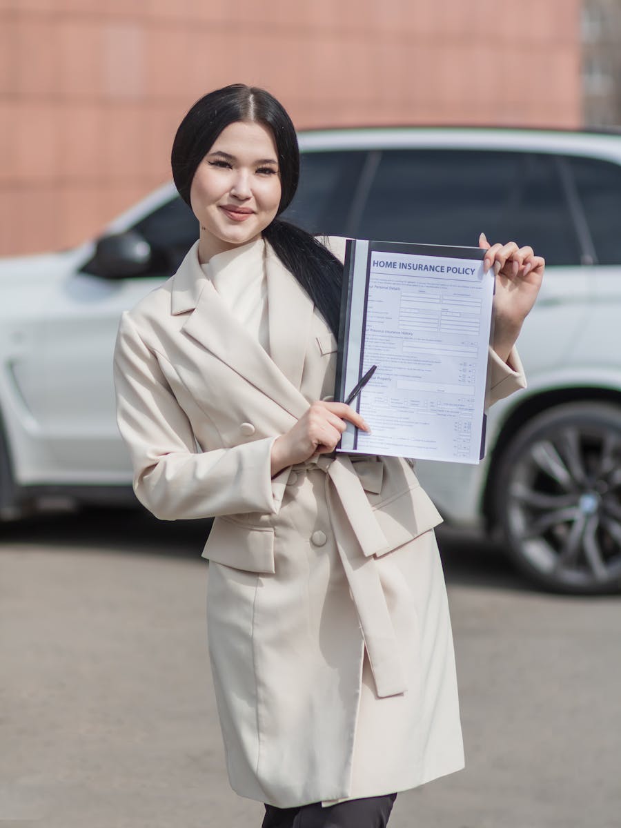 Smiling woman in beige blazer holding a home insurance policy document in outdoor setting.